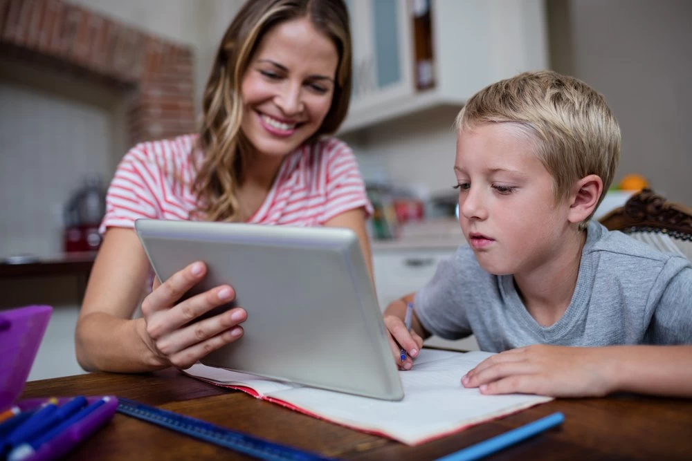 A homeschooling mum and son sitting at a desk. The mum is smiling and showing her son something on a tablet device, which the boy is studying intently.