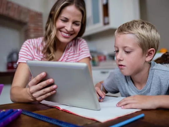 A homeschooling mum and son sitting at a desk. The mum is smiling and showing her son something on a tablet device, which the boy is studying intently.